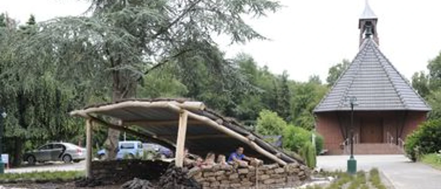 Kleine Holzhütte mit Menschen unter Dachkonstruktion, im Hintergrund steht eine Kapelle mit Turm.