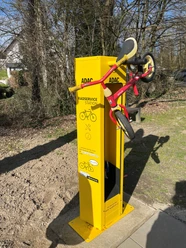 Radservice-Station Gelbe Radservice-Säule mit Fahrradaufhängung und Werkzeug, umgeben von Bäumen im Sonnenlicht.