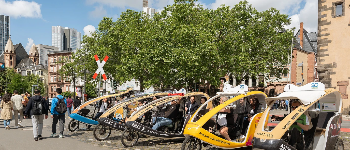 100 Jahre Neues Frankfurt – Die Velotaxi-Tour durch Bockenheim und das Westend Velotaxis warten in Frankfurt auf Fahrgäste, Menschen flanieren bei Sonnenschein.