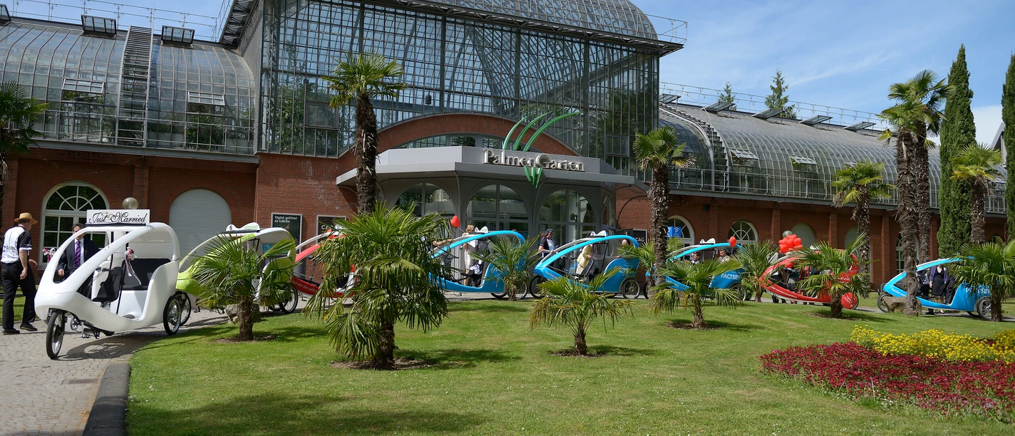 Frankfurt genussvoll erfahren Velotaxis vor dem Palmengarten Frankfurt, umgeben von Palmen und Glasfassade.