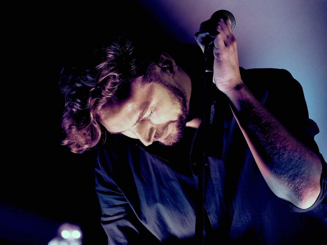 251129_FRaeuber_ReneGaens Musiker auf Bühne, im Lichtschein, hält Mikrofon. Intensiver Gesichtsausdruck und gesenkte Haltung.Musician on stage, in the light, holding microphone. Intense facial expression and lowered posture.Hudebník na pódiu, ve světle, s mikrofonem. Intenzivní výraz tváře a snížený postoj.Muzyk na scenie, w świetle, trzymający mikrofon. Intensywny wyraz twarzy i obniżona postawa.Muzikant op het podium, in het licht, met microfoon. Intense gezichtsuitdrukking en verlaagde houding.Musicista sul palco, sotto la luce, con il microfono in mano. Espressione facciale intensa e postura abbassata.