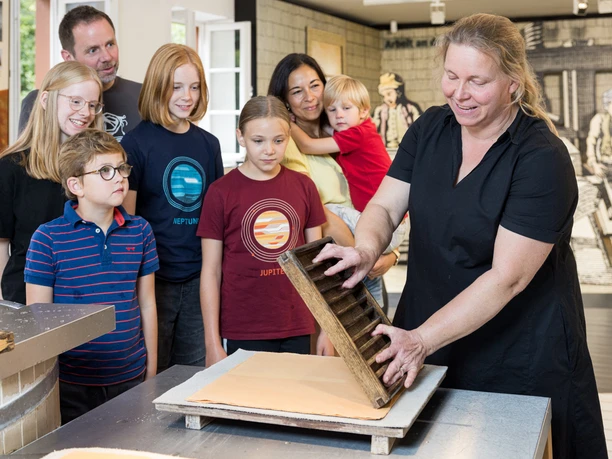 Papier – vom Handwerk zum Massenprodukt Gruppe von acht Personen, konzentriert um eine Frau, die traditionelles Papier schöpft.