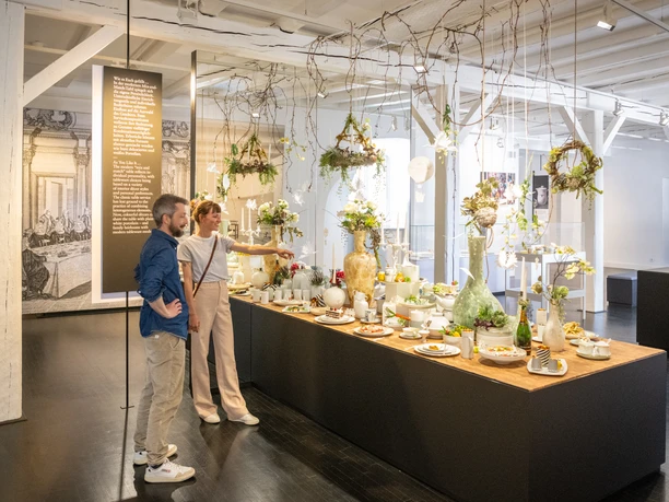 Zwei Personen betrachten im Fürstenberg-Museum dekorierte Tafel mit Porzellan und Blumengestecken.