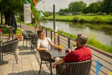 Menschen genießen Getränke auf einer Terrasse am Flussufer in Höxter, mit bunten Wimpeln dekoriert.