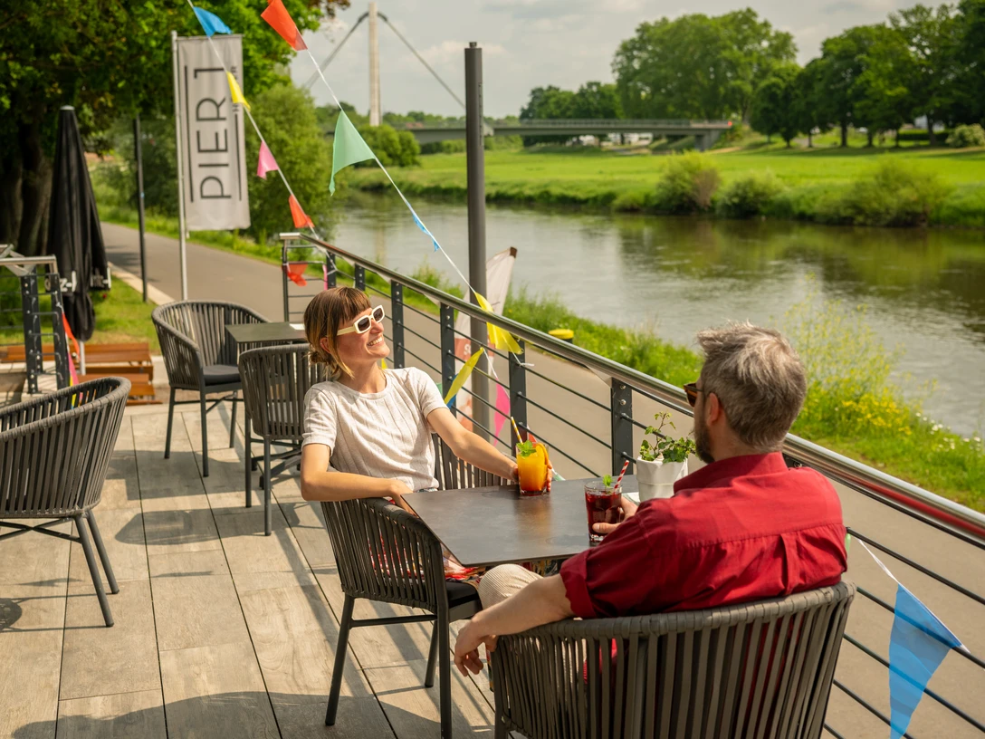 Hoexter-Gastronomie-Teutoburger-Wald-Stadt-Hoexter-D-Ketz-080.jpg Menschen genießen Getränke auf einer Terrasse am Flussufer in Höxter, mit bunten Wimpeln dekoriert.