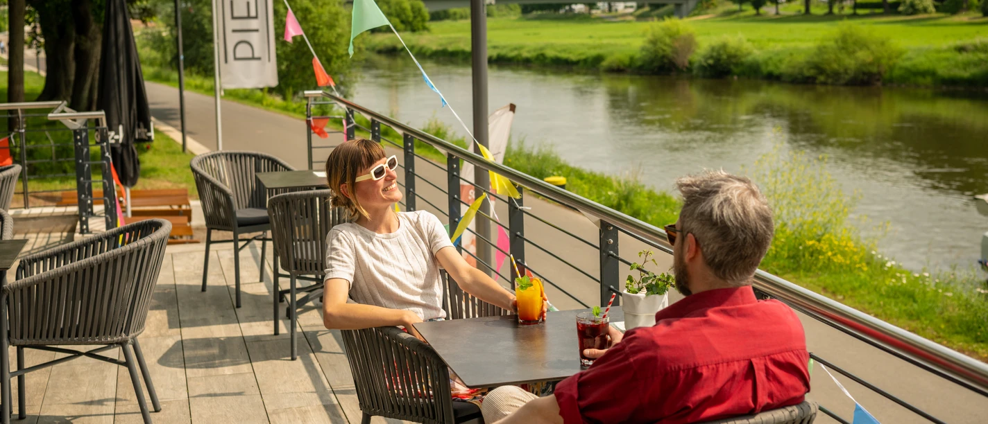 Hoexter-Gastronomie-Teutoburger-Wald-Stadt-Hoexter-D-Ketz-080.jpg Menschen genießen Getränke auf einer Terrasse am Flussufer in Höxter, mit bunten Wimpeln dekoriert.