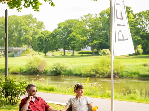 Hoexter-Gastronomie-Teutoburger-Wald-Stadt-Hoexter-D-Ketz-089.jpg Ein Paar sitzt entspannt auf Lounge-Möbeln in einem grünen Park in Höxter, nahe einem Fluss.