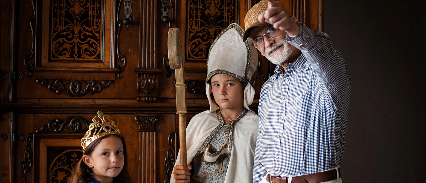 Online-Schloss-Kinderfuehrung-13.jpg Børn i middelalderkostumer og en ældre mand med hat i et historisk rum.