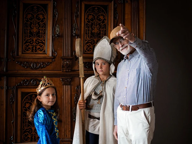 Online-Schloss-Kinderfuehrung-13.jpg Kinder in mittelalterlichen Kostümen und ein älterer Mann mit Hut in einem historischen Raum.
