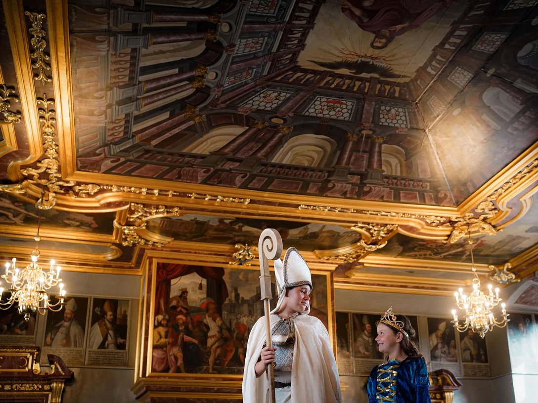 Online-Schloss-Kinderfuehrung-22.jpg Mittelalterlich kostümierter Bischof und Prinzessin in vergoldetem Barockraum unter prächtiger Decke.Bishop and princess in medieval costume in a gilded baroque room under a magnificent ceiling.Biskop og prinsesse i middelalderkostume i et forgyldt barokrum under et storslået loft.Bisschop en prinses in middeleeuws kostuum in een vergulde barokke kamer onder een prachtig plafond.