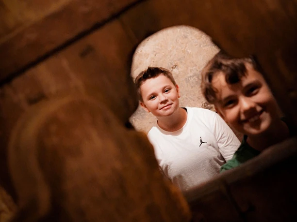 Online-Schloss-Kinderfuehrung-26.jpg To drenge kigger gennem en rustik trækonstruktion og griner til kameraet.