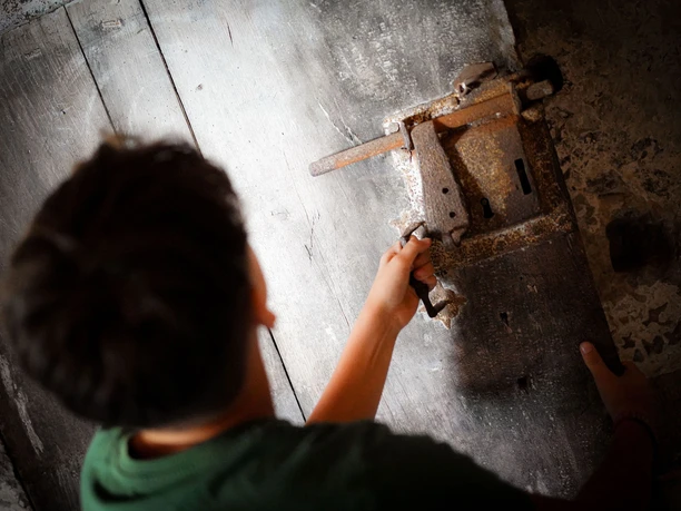 Online-Schloss-Kinderfuehrung-32.jpg Mand drejer et rustikt metalhåndtag på en trædør set ovenfra.