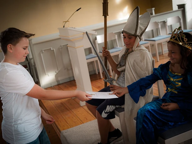 Online-Schloss-Kinderfuehrung-47.jpg Kinder in Mittelalterkostümen, ein Junge reicht einem sitzenden König ein Dokument.