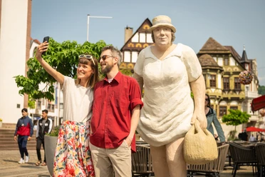 Frau und Mann machen Selfie neben großer Skulptur einer Frau. Im Hintergrund historische Gebäude.