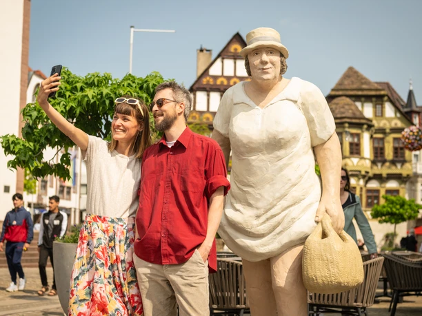 Frau und Mann machen Selfie neben großer Skulptur einer Frau. Im Hintergrund historische Gebäude.