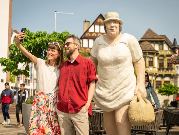 Frau und Mann machen Selfie neben großer Skulptur einer Frau. Im Hintergrund historische Gebäude.
