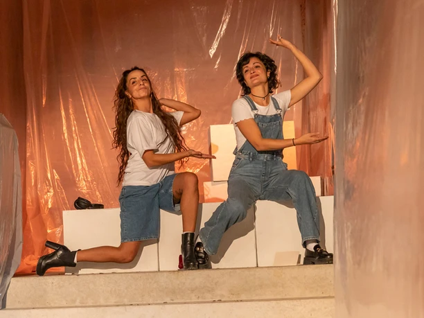 c.t.201 //in liebe, Zwei junge Frauen in Denim-Kleidung posieren theatralisch auf einer Bühne, eingehüllt in warmes Licht.
