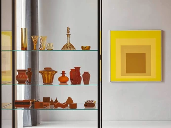 Josef Albers Hommage an das Quadrat – Gelb 1962, Öl auf Masonit Glasobjekte in warmen Brauntönen auf Glasregalen, Bild in gelber Farbskala rechts an der Wand.Glass objects in warm brown tones on glass shelves, picture in yellow color scale on the right wall.
