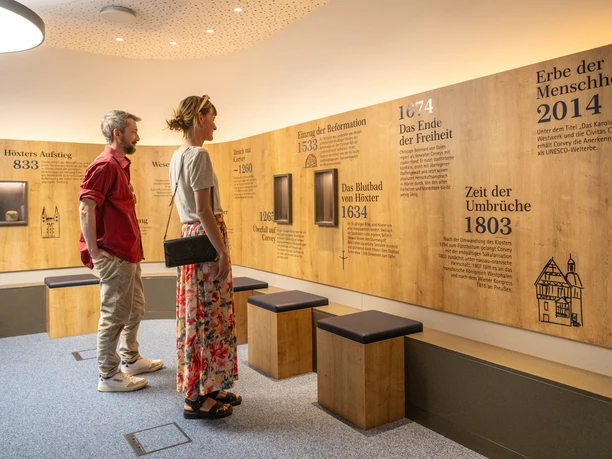 Zwei Personen erkunden eine Ausstellung zur Stadtgeschichte Höxters mit Infotafeln und Sitzgelegenheiten.