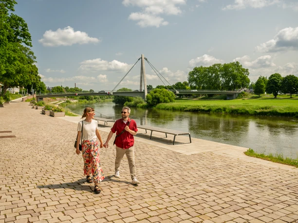 Zwei Personen spazieren entlang einer gepflasterten Promenade an der Weser, mit Brücke und grünem Ufer.
