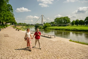 Hoexter-Weserpromenade-Teutoburger-Wald-Stadt-Hoexter-D-Ketz-095.jpg Zwei Personen spazieren entlang einer gepflasterten Promenade an der Weser, mit Brücke und grünem Ufer.