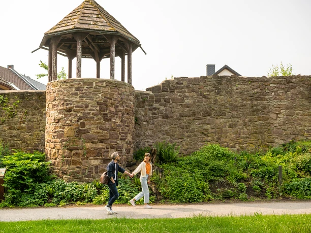 Hoexter-Stadtmauer-Teutoburger-Wald-Stadt-Hoexter-D-Ketz-048.jpg Zwei Frauen spazieren an einer historischen Stadtmauer in Höxter vorbei, umgeben von Grün.