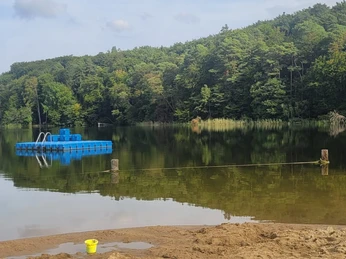 Sandstrand und Badeinsel - Badestelle Campingplatz Lütauer See.jpeg Sandstrand mit gelbem Eimer, ruhigem See, grüne Uferbäumen und einer blauen Badeinsel im Wasser.