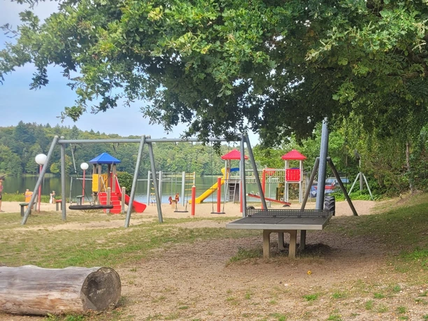 Spielplatz mit Klettergerüst und Rutsche am Lütauer See, umgeben von Bäumen.
