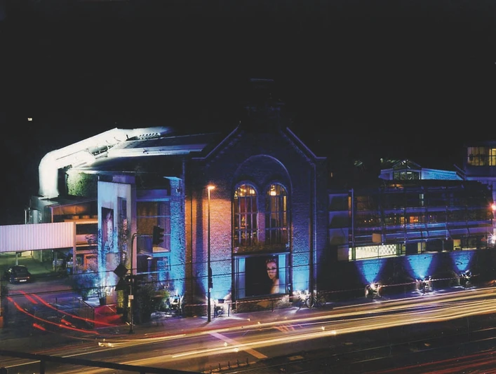 BALLONI Halle_1 Alte Backsteinhalle bei Nacht, beleuchtet in Blau, mit vorbeiziehenden Scheinwerferlichtern.Old brick hall at night, illuminated in blue, with passing spotlights.