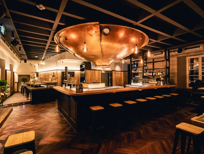 Gastraum im Bürgerhaus Ratingen. Ein zentraler Raum mit einem großen kupferfarbenen Kessel an der Decke, umgeben von Holzmöbeln.