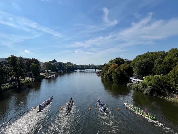 Vier Drachenboot-Teams in rasanter Fahrt auf einem Fluss. Bäume säumen das Ufer.