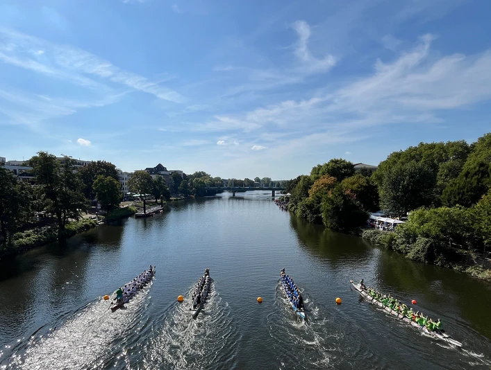 Drachenboot-Festival, Mülheim an der Ruhr Vier Drachenboot-Teams in rasanter Fahrt auf einem Fluss. Bäume säumen das Ufer.