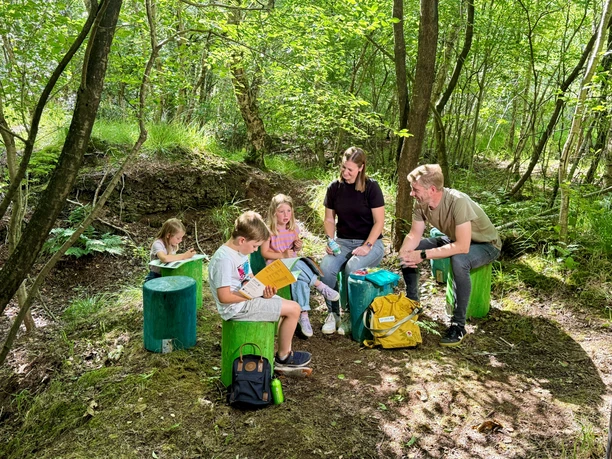 GruenesKlassenzimmer_am_Bodenprofil_Provinzialmoor_VS_2.jpg Kinder und Erwachsene sitzen im Wald auf grünen Holzhockern und lernen gemeinsam im Freien.