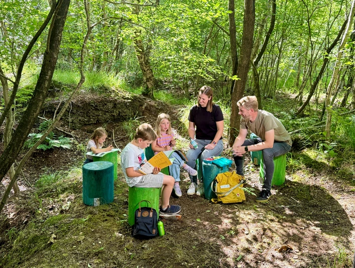 GruenesKlassenzimmer_am_Bodenprofil_Provinzialmoor_VS_2.jpg Kinder und Erwachsene sitzen im Wald auf grünen Holzhockern und lernen gemeinsam im Freien.