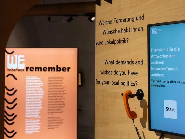 Blick in die Ausstellung Eine moderne Ausstellungseinheit mit interaktivem Bildschirm und orangefarbenem Telefon, umgeben von thematischen Plakaten.