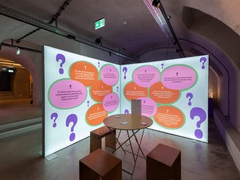 View of the exhibition Ausstellungsraum mit beleuchteten Tafeln und modernen Sitzwürfeln für Besucher.Exhibition room with illuminated panels and modern seating cubes for visitors.