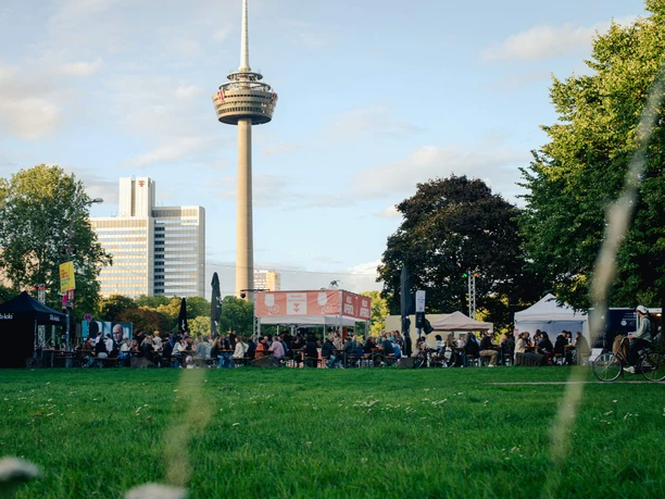 Apéro Markt in Köln Ein belebter Markt auf einer grünen Wiese, umgeben von Bäumen und dem markanten Colonius-Turm im Hintergrund.