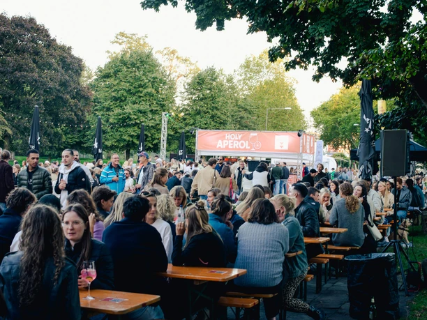 Apéro Markt in Köln Menschen genießen an Tischen Getränke und Unterhaltung im Freien, umgeben von Bäumen.