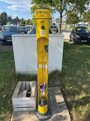 Radservice-Station Das Bild zeigt eine gelbe Radservice-Station mit Werkzeugen, umgeben von geparkten Autos und Bäumen.