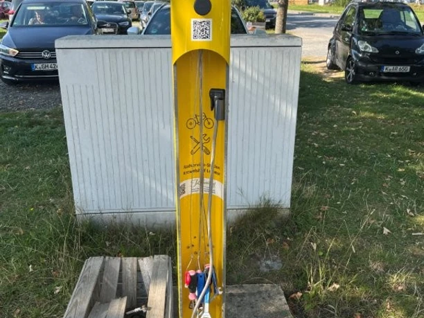 Radservice-Station Das Bild zeigt eine gelbe Radservice-Station mit Werkzeugen, umgeben von geparkten Autos und Bäumen.