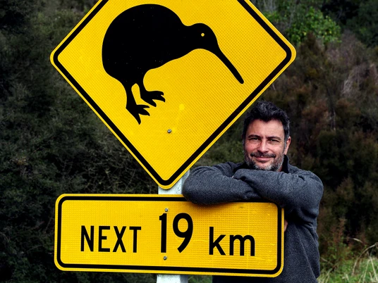 Pressebild Neuseeland - Kiwi-Schild Ein Mann steht angelehnt hinter einem Schild mit zwei Schildern; auf dem oberen ist ein Kiwi Vogel zu sehen, auf dem unteren Schild steht 19km