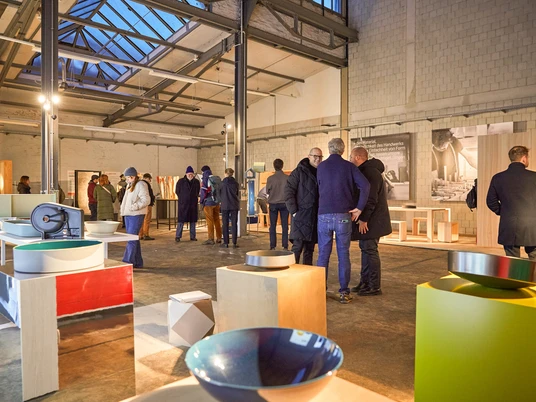 MACHWERKHAUS Innenansicht Ausstellungshalle mit modernen Designobjekten und Besuchergruppen im Gespräch.Exhibition hall with modern design objects and groups of visitors in conversation.