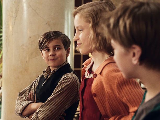 Filmstill zu "Das geheime Stockwerk" Drei Kinder stehen in einem hellen Raum, sie lächeln einander freundlich an.