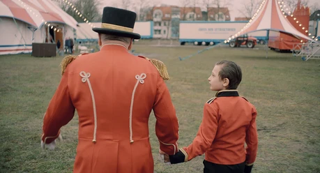 Film still for "Zirkuskind" A ringmaster and a child in a red uniform walk hand in hand onto a circus meadow.