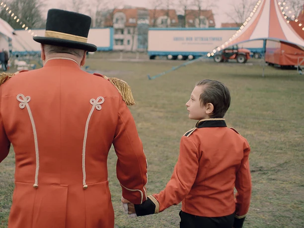Film still for "Zirkuskind" A ringmaster and a child in a red uniform walk hand in hand onto a circus meadow.