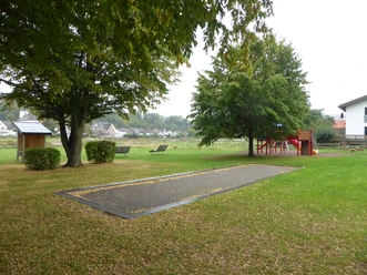 Eine Boulebahn auf einer Wiese mit Bäumen und einem Spielplatz im Hintergrund..