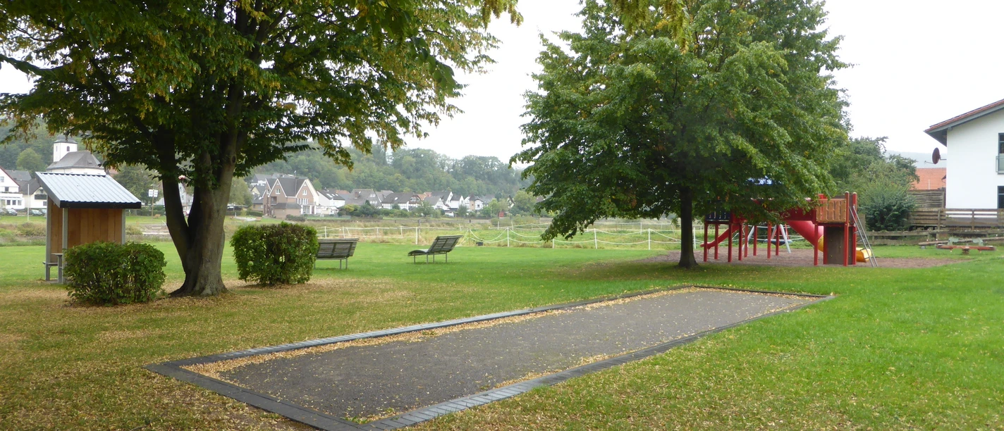Eine Boulebahn auf einer Wiese mit Bäumen und einem Spielplatz im Hintergrund..