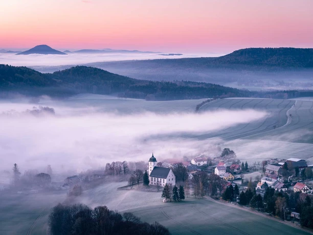 View to Papstdorf from the Papststein Fog-covered landscape at sunrise with church and Papstdorf in the foreground, hills in the background.