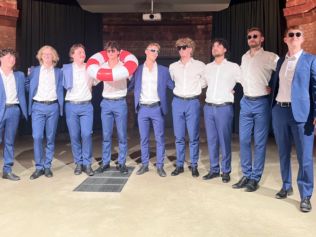 twentytwo-ensemble-(C)-RX-1920x1080px.jpeg Neun Männer in blauen Anzügen und Sonnenbrillen posieren in einer Backsteinhalle.