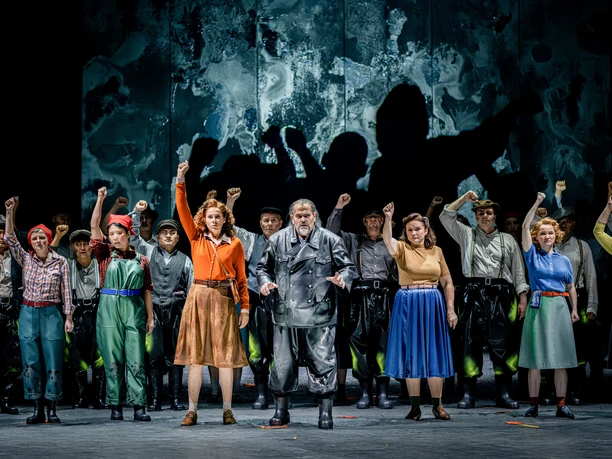 Eine Theatergruppe auf der Bühne hebt Arme im Jubel. Tänzer in farbenfrohen Outfits.